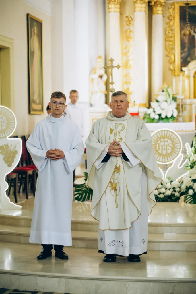 Chłopiec w stroju komunijnym stoi obok księdza
